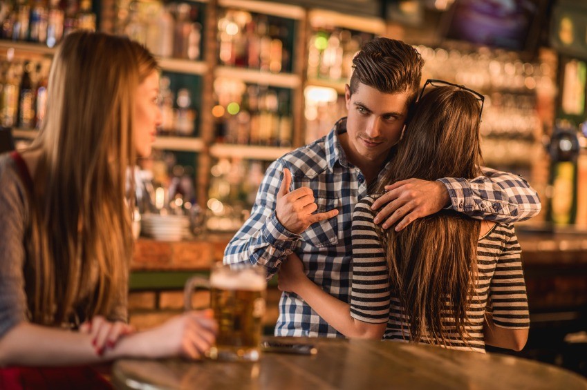 Unfaithful young man in a cafe. He is embracing his girlfriend and making call gesture to another woman behind her back.